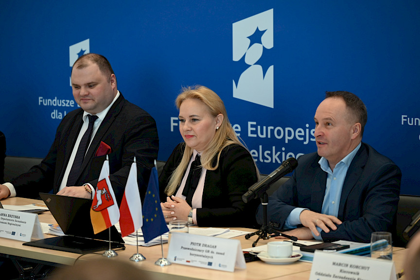 XVII Posiedzenie grup roboczych działających przy Komitecie Monitorującym Fundusze Europejskie dla Lubelskiego 2021-2027, trójka prezydium siedząca za stołem, w tle ścianka Funduszy Europejskich.