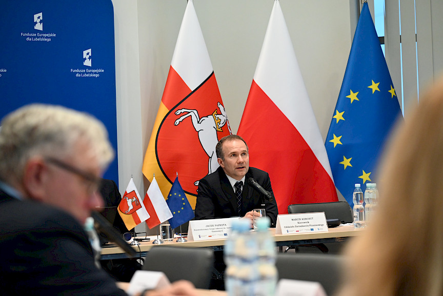 XVII Posiedzenie grup roboczych działających przy Komitecie Monitorującym Fundusze Europejskie dla Lubelskiego 2021-2027, na pierwszym planie członek prezydium, w tle flagi.