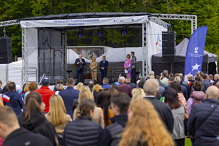 Zdjęcie wspólne XII finału Dni Funduszy Europejskich. Scena, na scenie Marszałek Jarosław Stawiarski z osobami towarzyszącymi. Przed sceną ludzie siedzący na ławkach, kobiety, mężczyźni, dzieci. Marszałek przemawia do zgromadzonych osób.