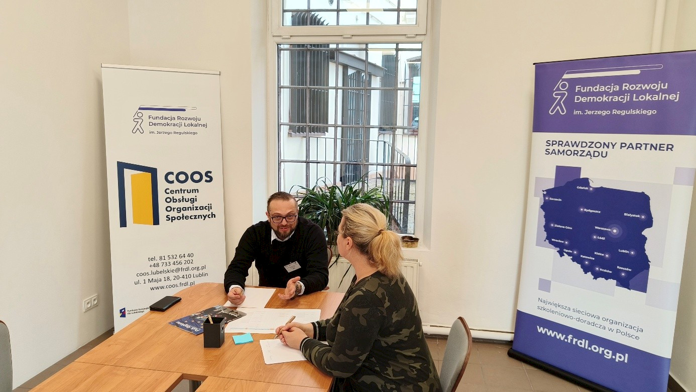 Biały pokój z oknem. W oknie kraty, w pokoju stół o fakturze drewna. Przy stole mężczyzna i kobieta. Rozmawiają. Za nimi rollupy