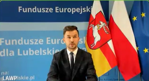 Mężczyzna w garniturze przemawia na tle flag Polski, Unii Europejskiej i województwa lubelskiego przed tablicą z napisem „Fundusze Europejskie dla Lubelskiego
