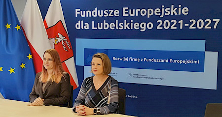Kobiety siedzą przy stole, za nimi ścianka z napisem: „Fundusze Europejskie dla Lubelskiego 2021”, obok trzy flagi: województwa lubelskiego, Rzeczpospolitej Polski, Unii Europejskiej.