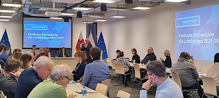 Grupa kobiet i mężczyzn siedzi na krzesłach, zwróconych w stronę kobiety stojącej przed nimi z mikrofonem w dłoni. Za prowadzącym znajdują się trzy flagi: Województwa Lubelskiego, Rzeczypospolitej Polskiej oraz Unii Europejskiej. W tle widoczny jest ekran