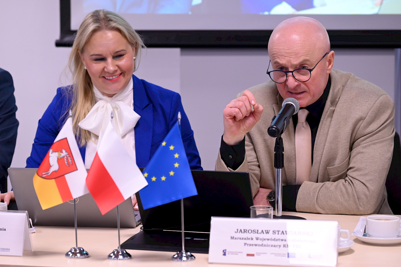 Marszałek Jarosław Stawiarski wypowiada się do mikrofonu. Po lewej stronie zdjęcia obok niego siedzi uśmiechnięta Dyrektor Anna Brzyska,