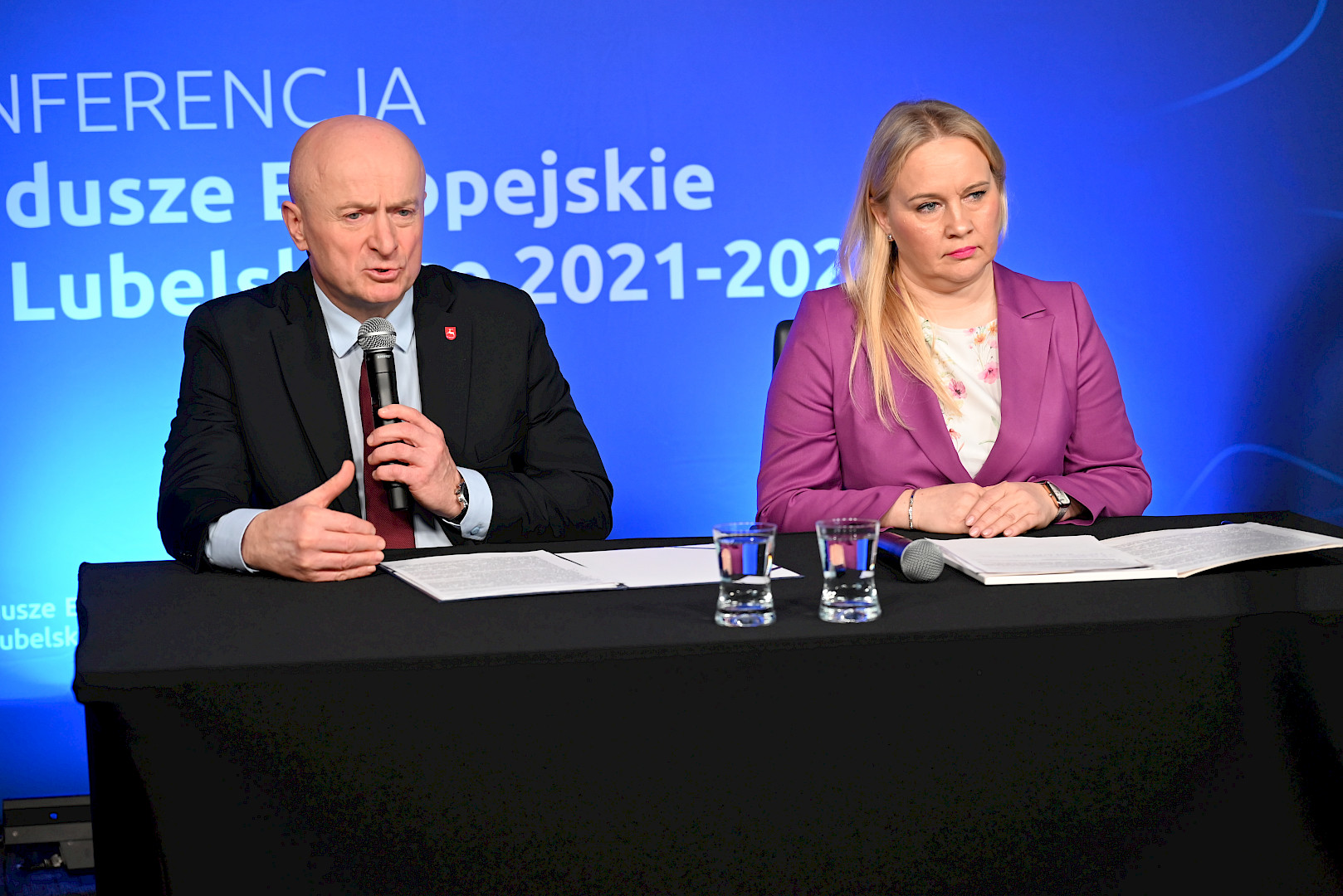 Marszałek Województwa Lubelskiego Jarosław Stawiarski siedzi za stołem i wypowiada się do mikrofonu. Po jego lewej stronie siedzi Dyrektor Departamentu Zarządzania Programami Regionalnymi Anna Brzyska.