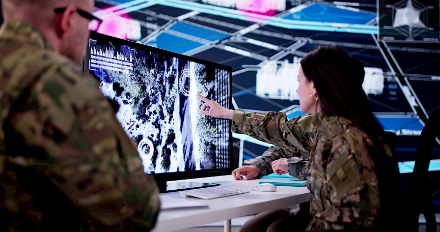 Zdjęcie przedstawia kobietę w wojskowym mundurze wskazującą palcem na monitor komputera. Po lewej stronie stoi mężczyzna w wojskowym mundurze patrzący w stronę ekranu.