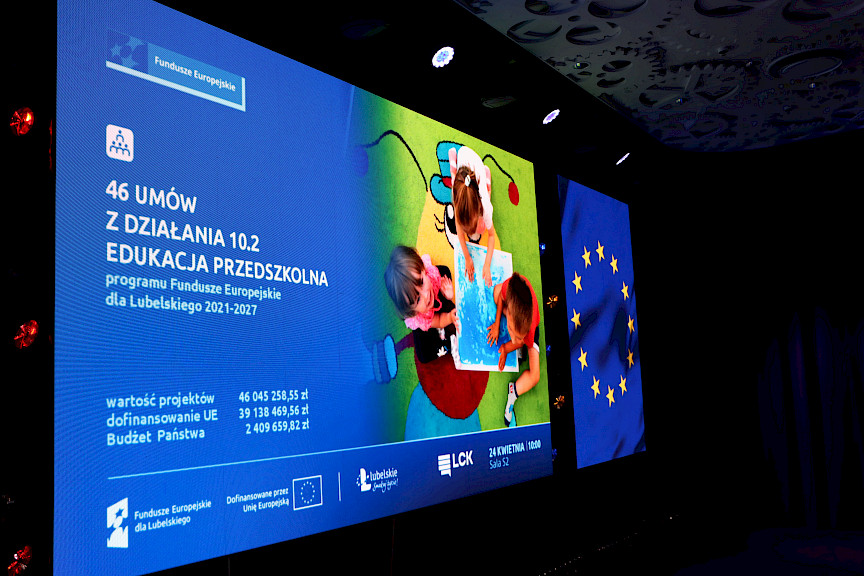 Ujęcie tablicy led na ścianie z wyświetlonymi informacjami na temat wydarzenia. Po prawej stronie wyświetlacza widnieją dzieci przy przedszkolnym stoliku patrzące do góry.