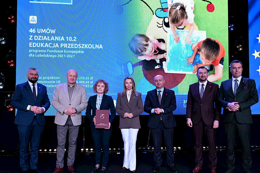 Zarząd Województwa Lubelskiego pozuje do zdjęcia z beneficjentką projektu. Kobieta trzyma teczkę w umową.