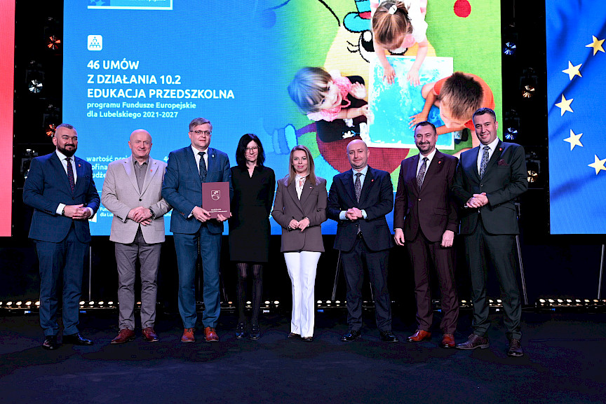 Zarząd Województwa Lubelskiego pozuje do zdjęcia z beneficjentami projektu. Mężczyzna trzyma teczkę w umową.