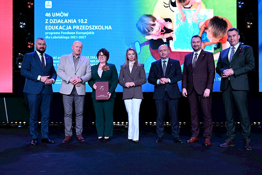 Zarząd Województwa Lubelskiego pozuje do zdjęcia z beneficjentką projektu. Kobieta trzyma teczkę w umową.