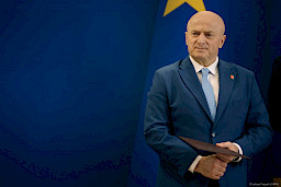 Marszałka Województwa Lubelskiego Jarosława Stawiarskiego trzymający bordową teczkę z umową czeka na podejście beneficjenta.