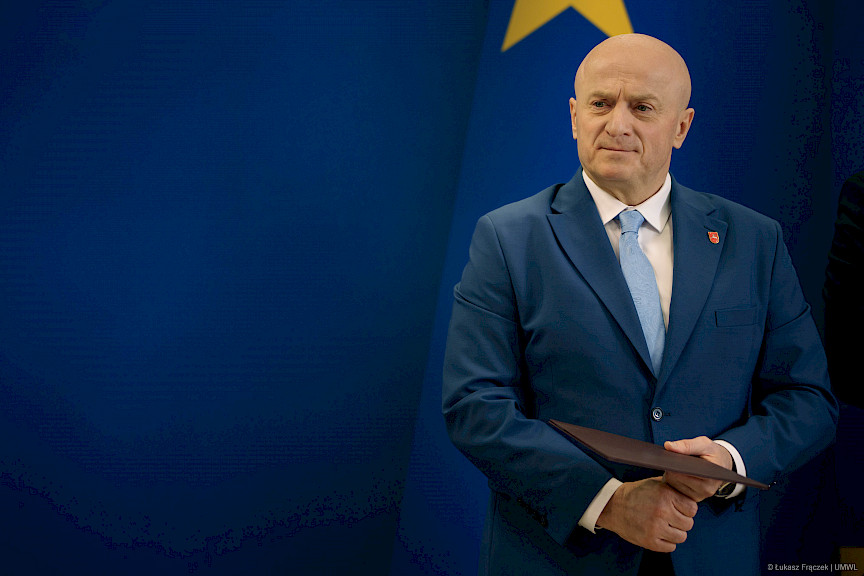 Marszałka Województwa Lubelskiego Jarosława Stawiarskiego trzymający bordową teczkę z umową czeka na podejście beneficjenta.