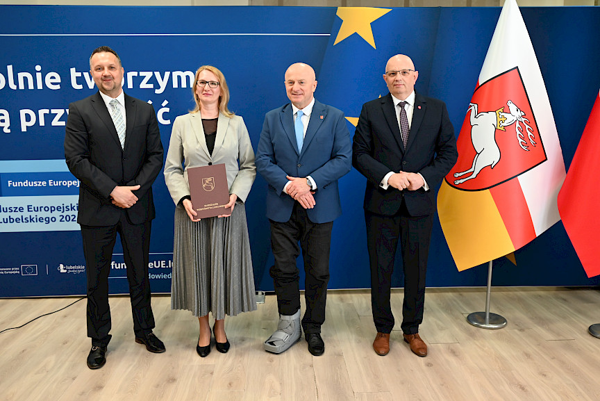 Marszałek Jarosław Stawiarski pozuje do zdjęć z Dyrektorem Wojewódzkiego Urzędu Pracy w Lublinie Piotrem Pitucha oraz Beneficjentką projektu trzymającą teczkę z umową. Obok kobiety stoi mężczyzna.