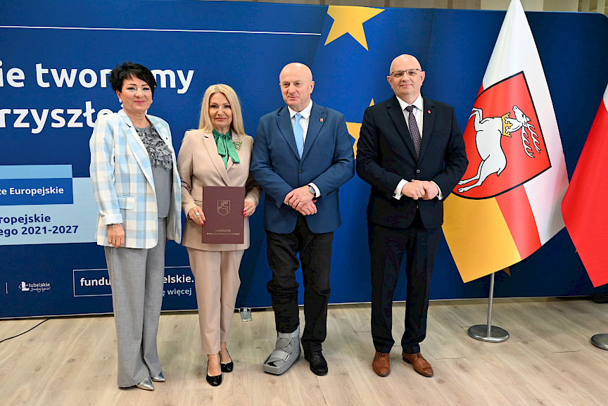 Marszałek Jarosław Stawiarski pozuje do zdjęć z Dyrektorem Wojewódzkiego Urzędu Pracy w Lublinie Piotrem Pitucha oraz Beneficjentką projektu trzymającą teczkę z umową. Obok kobiety stoi druga kobieta,