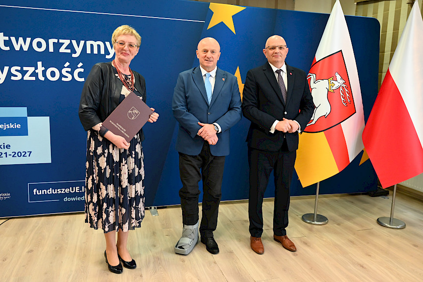 Marszałek Województwa Lubelskiego Jarosław Stawiarski pozuje do zdjęć z Beneficjentką projektu trzymającą teczkę z umową. Obok nich stoi Dyrektor Wojewódzkiego Urzędu Pracy Tomasz Pitucha.