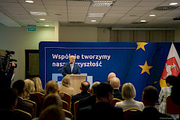 Marszałek Województwa Lubelskiego Jarosław Stawiarski przemawia do mikrofonu do uczestników spotkania siedzących na widowni.  Za nim znajduje się ścianka w kolorze granatowym z napisami nawiązującymi do Funduszy Europejskich.
