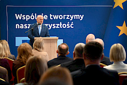Marszałek Województwa Lubelskiego Jarosław Stawiarski przemawia do mikrofonu do uczestników spotkania siedzących na widowni.  Za nim znajduje się ścianka w kolorze granatowym z napisami nawiązującymi do Funduszy Europejskich.