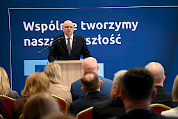 Dyrektor Wojewódzkiego Urzędu w Lublinie Pracy Tomasz Pitucha. przemawia do mikrofonu do uczestników spotkania siedzących na widowni.  Za nim znajduje się ścianka w kolorze granatowym z napisami nawiązującymi do Funduszy Europejskich.