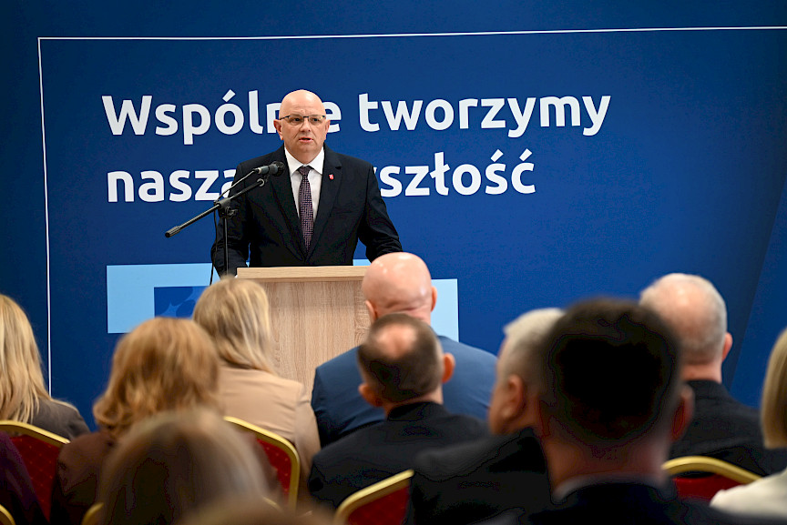 Dyrektor Wojewódzkiego Urzędu w Lublinie Pracy Tomasz Pitucha. przemawia do mikrofonu do uczestników spotkania siedzących na widowni.  Za nim znajduje się ścianka w kolorze granatowym z napisami nawiązującymi do Funduszy Europejskich.