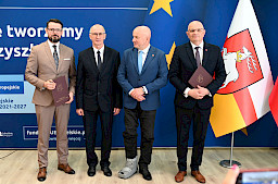 Marszałek Województwa Lubelskiego pozuje do zdjęć z dwoma mężczyznami, beneficjentami projektu, jeden z nich trzyma teczkę z umową. Obok Marszałka stoi dyrektor Wojewódzkiego Urzędu Pracy w Lublinie Tomasz Pitucha.