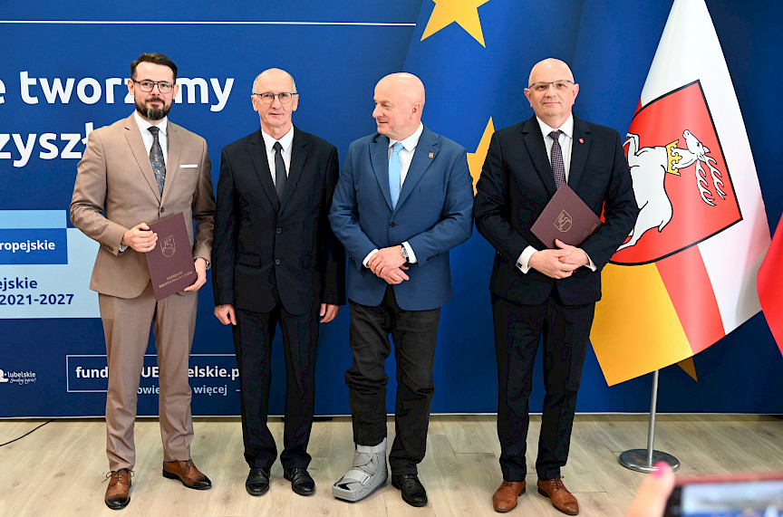 Marszałek Województwa Lubelskiego pozuje do zdjęć z dwoma mężczyznami, beneficjentami projektu, jeden z nich trzyma teczkę z umową. Obok Marszałka stoi dyrektor Wojewódzkiego Urzędu Pracy w Lublinie Tomasz Pitucha.
