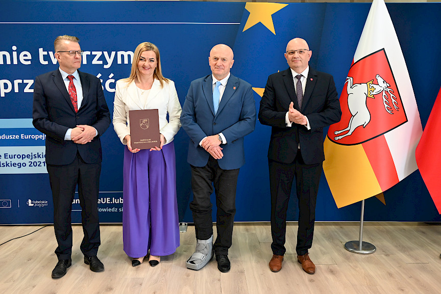 Marszałek Województwa Lubelskiego pozuje do zdjęć z beneficjentami projektu, mężczyzną oraz kobietą trzymającą teczkę z umową. Obok Marszałka stoi dyrektor Wojewódzkiego Urzędu Pracy w Lublinie Tomasz Pitucha.