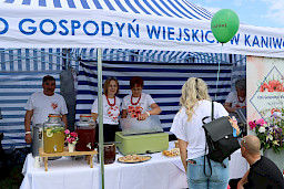 Dwie kobiety oraz mężczyzna stoją pod namiotem koła gospodyń wiejskich. Na stole stoją słoje z lemoniadą oraz pojemniki z jedzeniem.