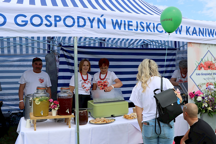 Dwie kobiety oraz mężczyzna stoją pod namiotem koła gospodyń wiejskich. Na stole stoją słoje z lemoniadą oraz pojemniki z jedzeniem.