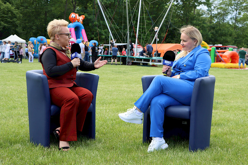 Dyrektor Departamentu Zarządzania Programami Regionalnymi Anna Brzyska udziela wywiadu do telewizji. Wraz z dziennikarką siedzą na niebieskich fotelach ustawionych na terenie wydarzenia. W tle widoczne są atrakcje dla dzieci.