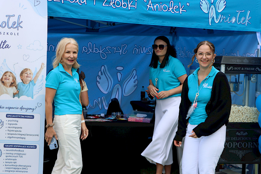 Trzy kobiety, przedstawicielki żłobka stoją przy swoim niebieskim namiocie i  uśmiechają się do zdjęcia.