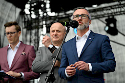 Burmistrz Miasta Łęczna przemawia do mikrofonu ze sceny. Obok niego stoi Marszałek Jarosław Stawiarski oraz dalej mężczyzna, który prowadzi wydarzenie na scenie.