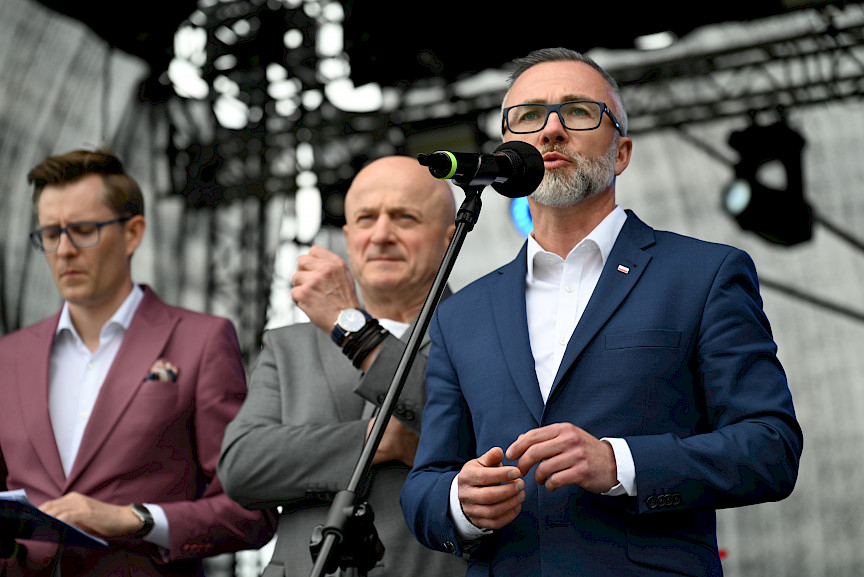 Burmistrz Miasta Łęczna przemawia do mikrofonu ze sceny. Obok niego stoi Marszałek Jarosław Stawiarski oraz dalej mężczyzna, który prowadzi wydarzenie na scenie.