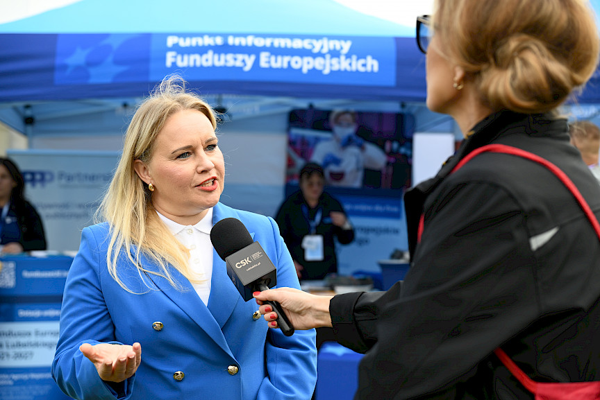 Dyrektor Departamentu Zarządzania Programami Regionalnymi Anna Brzyska udziela wywiadu do mikrofonu. W tle znajduje się niebieski namiot ze stoiskiem punktu informacyjnego o Funduszach Europejskich.
