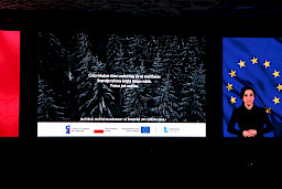 Na ekranie led wyświetlona jest informacja o uzależnieniu dzieci. Po prawej stronie wyświetlona jest flaga Unii Europejskiej oraz stoi kobieta tłumaczka języka migowego.