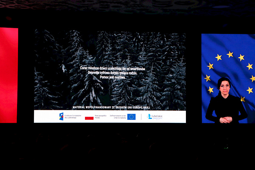 Na ekranie led wyświetlona jest informacja o uzależnieniu dzieci. Po prawej stronie wyświetlona jest flaga Unii Europejskiej oraz stoi kobieta tłumaczka języka migowego.