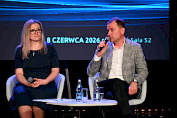 Kobieta oraz mężczyzna siedzą na krzesłach na scenie, mężczyzna wypowiada się do mikrofonu w ramach panelu dyskusyjnego.