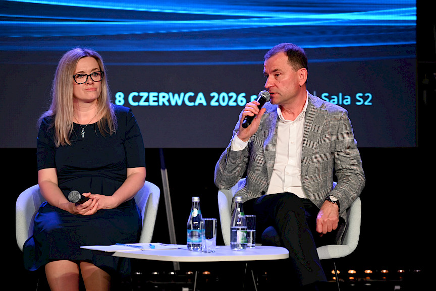 Kobieta oraz mężczyzna siedzą na krzesłach na scenie, mężczyzna wypowiada się do mikrofonu w ramach panelu dyskusyjnego.