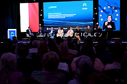 Ujęcie na cześć widowni od tyłu. Na scenie widoczni są uczestnicy panelu dyskusyjnego siedzący na krzesłach.