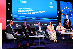 Trzy kobiety oraz trzech mężczyzn siedzi na krzesłach na scenie podczas panelu dyskusyjnego. Mężczyzna wypowiada się do mikrofonu.