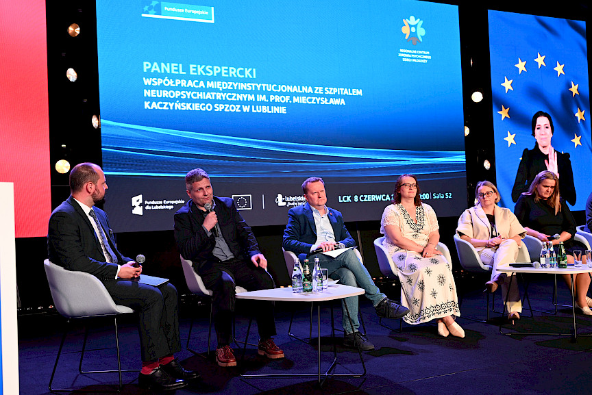 Trzy kobiety oraz trzech mężczyzn siedzi na krzesłach na scenie podczas panelu dyskusyjnego. Mężczyzna wypowiada się do mikrofonu.