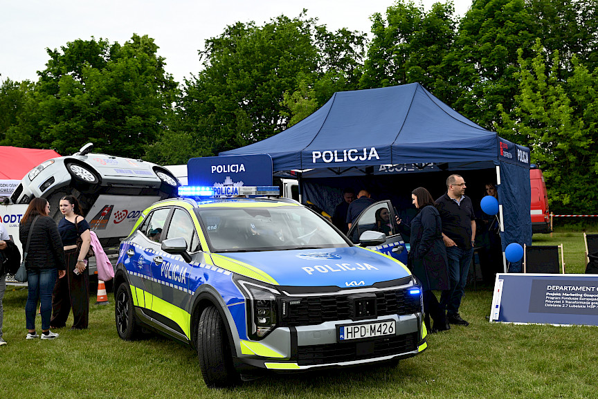 Stoisko policji, samochód policyjny, ludzie chodzący wokół i przyglądający się.