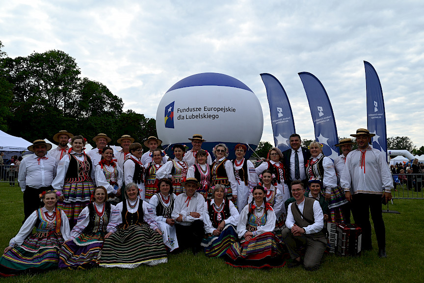Ludzie w strojach ludowych pozujący do zdjęcia na tle balonu z napisem Fundusze Europejskie dla Lubelskiego i winderów z logo.