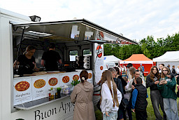 Food truck z pizzą, w środku pani przygotowująca jedzenie, obok ludzie czekający w kolejce