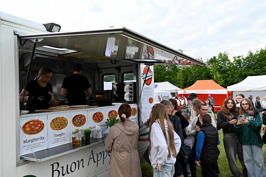 Food truck z pizzą, w środku pani przygotowująca jedzenie, obok ludzie czekający w kolejce