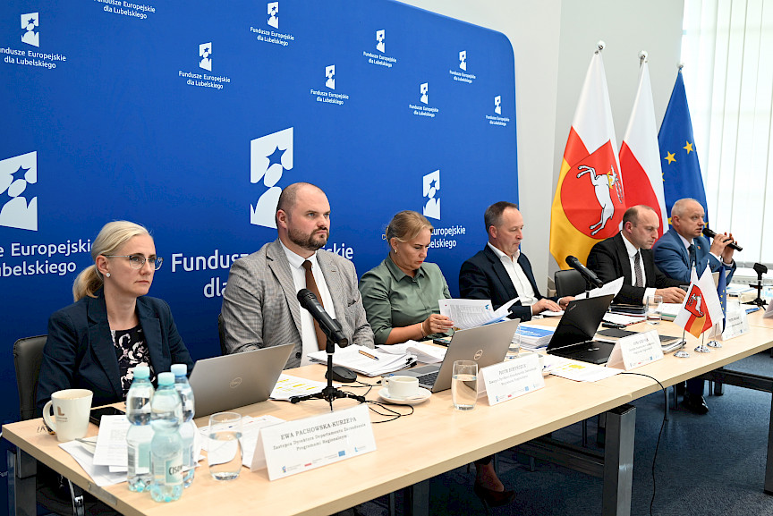 Dyrekcja Departamentu Zarządzania Programami Regionalnymi oraz przedstawiciele Departamentu Europejskiego Funduszu Rozwoju Regionalnego siedzący za stołem. Na stole znajdują się dokumenty, laptopy, mikrofonu, woda