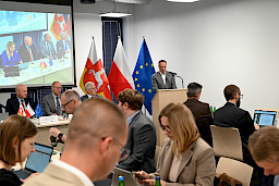 Mężczyzna wypowiada się przy ambonie do mikrofonu, widoczna jest również część uczestników posiedzenia siedzących na twarzy.