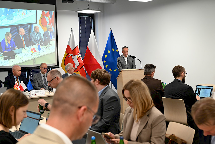 Mężczyzna wypowiada się przy ambonie do mikrofonu, widoczna jest również część uczestników posiedzenia siedzących na twarzy.