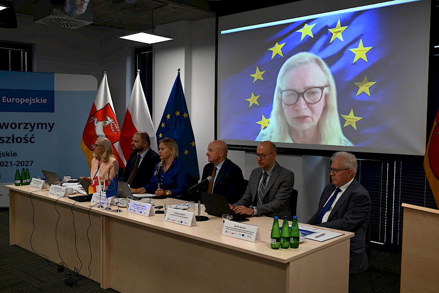 Przewodniczący komisji siedzą za stołem, za ich plecami wyświetlana jest twarz kobiety połączonej zdalnie na spotkaniu.
