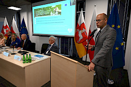 Przedstawiciel Komisji Europejskiej wypowiada się do mikrofonu za ambony. Wyświetla prezentację na ekranie rzutnika. Obok widać siedzących za stołem przewodniczących posiedzenia.