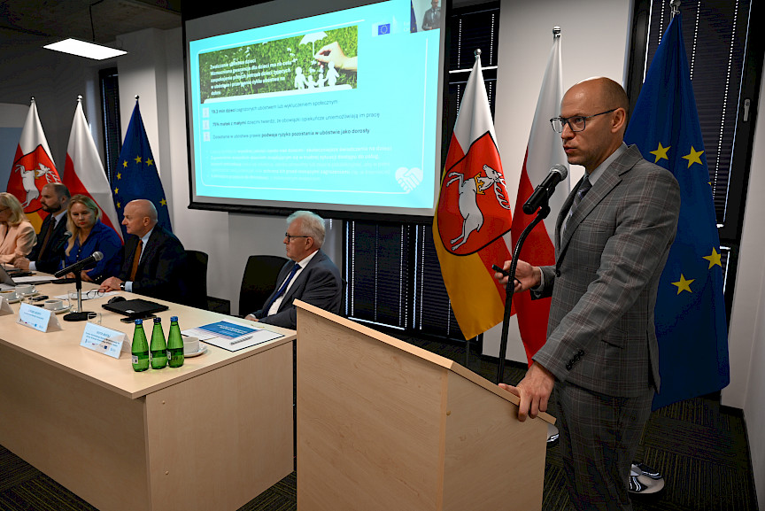 Przedstawiciel Komisji Europejskiej wypowiada się do mikrofonu za ambony. Wyświetla prezentację na ekranie rzutnika. Obok widać siedzących za stołem przewodniczących posiedzenia.